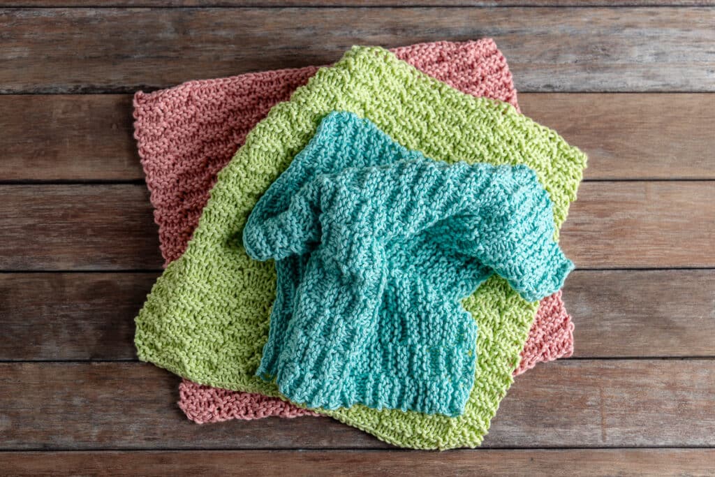 3 knitted dishcloths in pink, green, and blue on a wooden background