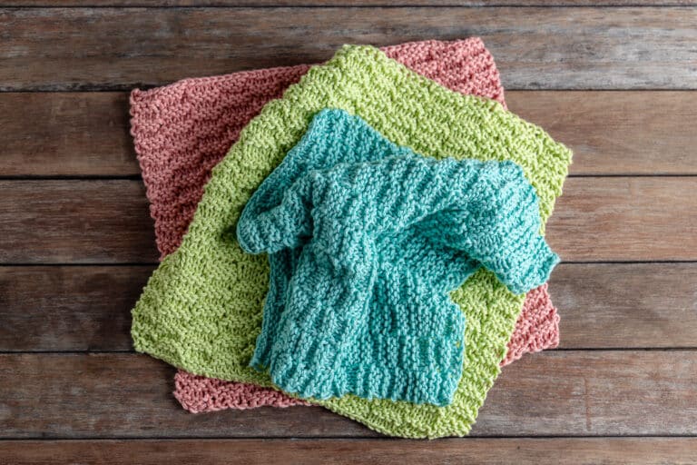 3 knitted dishcloths in pink, green, and blue on a wooden background