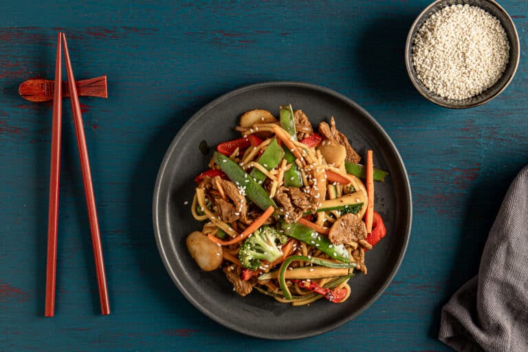 beef stir fry with lime and kekap manis with chop sticks, a napkin, and a bowl of sesame seeds on a dark background