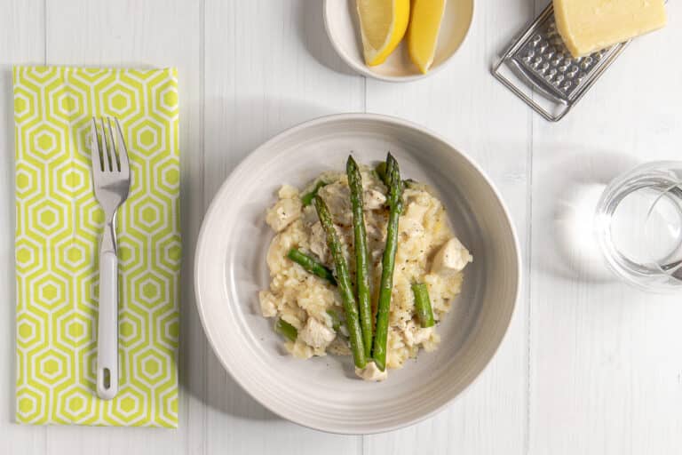 asparagus and chicken risotto in a bowl with a fork, napkin, water glass, lemon wedges, parmesan, grater