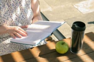 woman reading book on her lunch break at a city table with a reusable coffee cup and apple