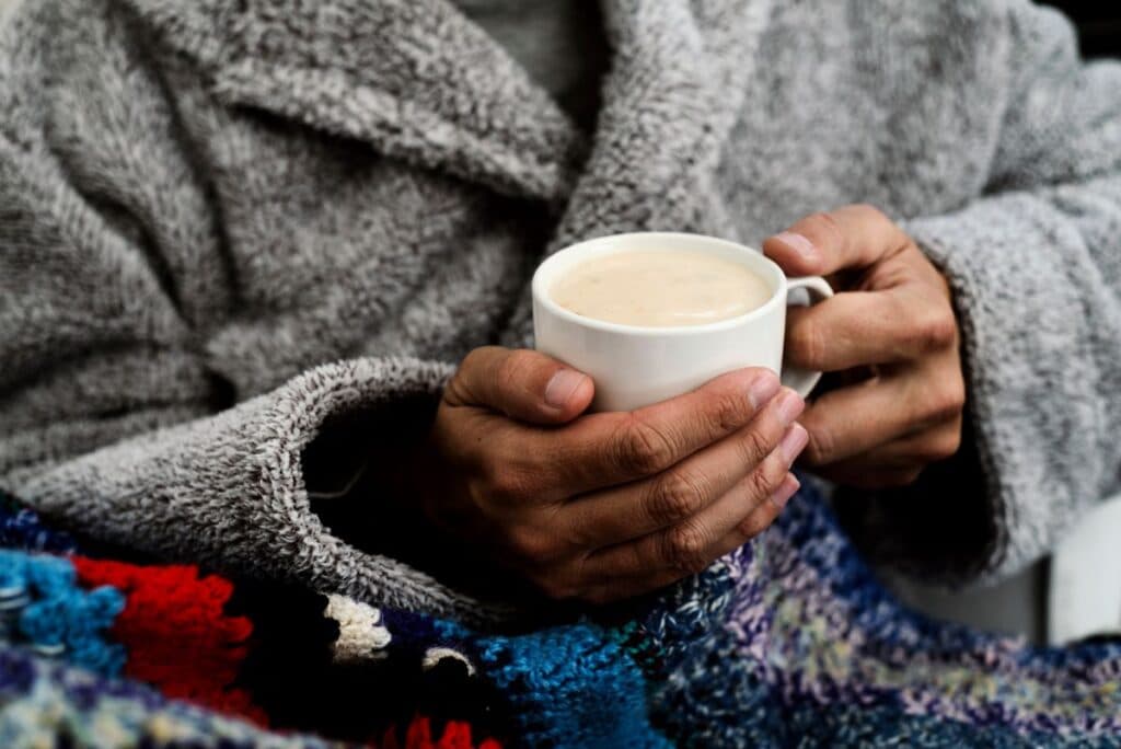 man wearing a warm robe and blanket and holding a hot drink to stay warm without heating