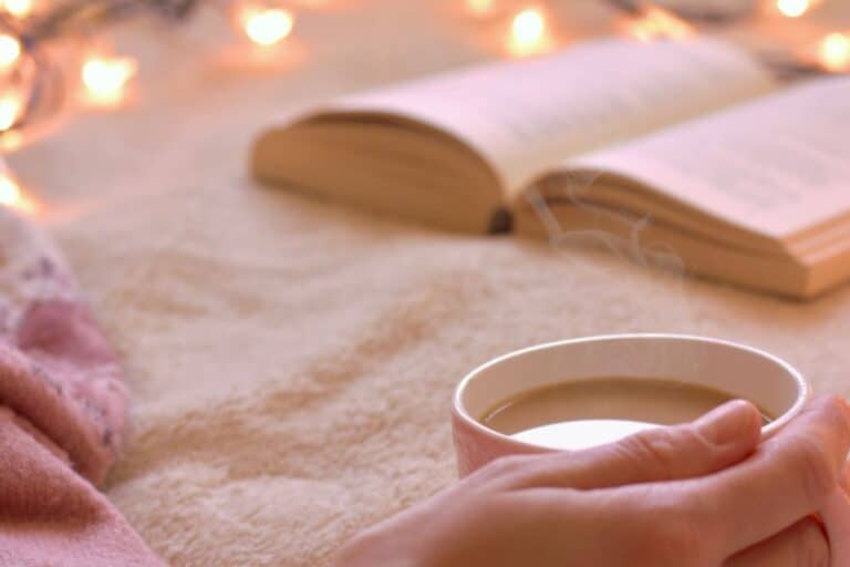 woman reading book in bed with coffee, blankets, fairy lights