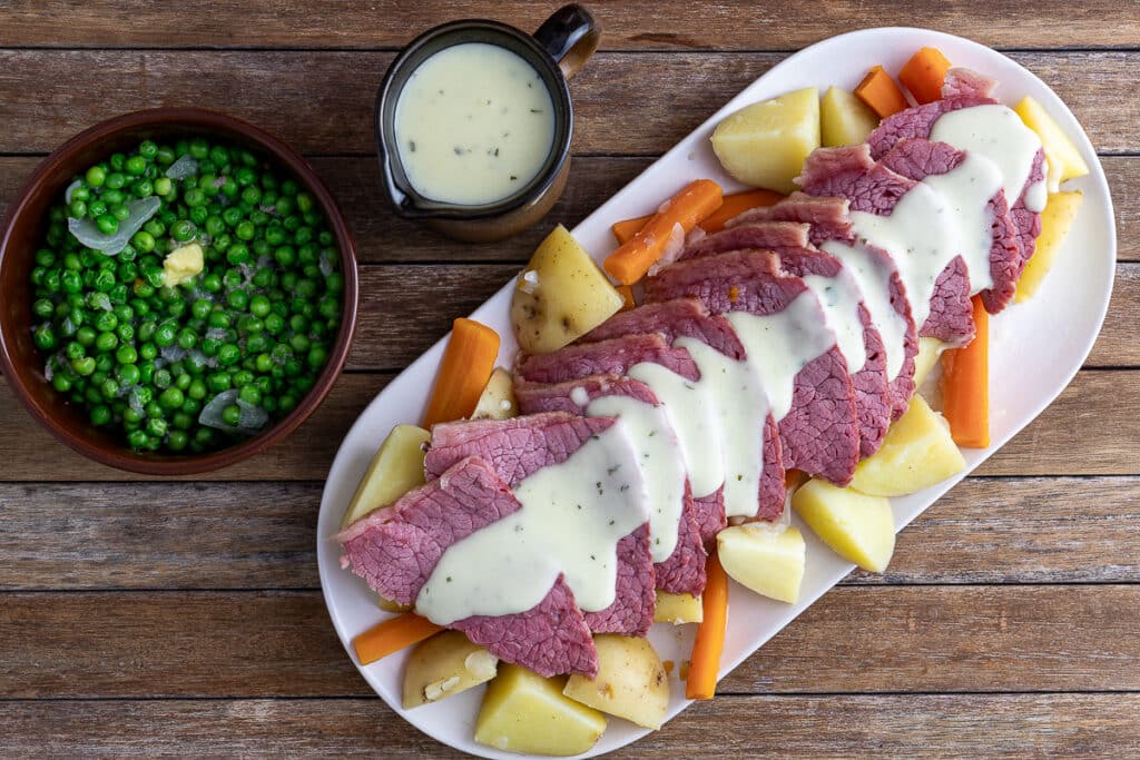 traditional corned beef on a platter with boiled potato and carrot and a side of peas and white sauce