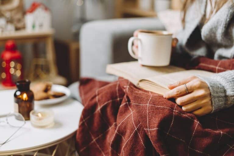woman reading with a blanket on her lap and a hot drink representing hygge on a budget